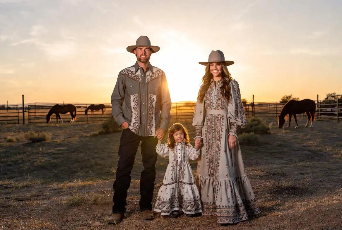 A family of three dressed in matching embroidered western-style clothing, featuring intricate patterns on a light gray fabric with long sleeves and flowing skirts or pants.